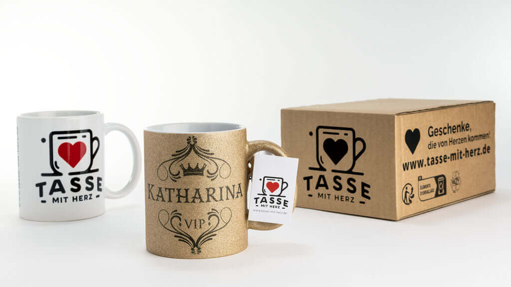 Goldene Glitzertasse mit VIP-Schriftzug und dem Namen Katharina, präsentiert neben einer weißen Tasse mit Herz-Logo und einem Versandkarton mit der Aufschrift ‚Geschenke, die von Herzen kommen!‘ von Tasse mit Herz.